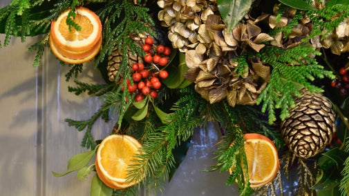 A Christmas wreath with gold flowers, oranges and red berries intertwined in foliage hangs on a door